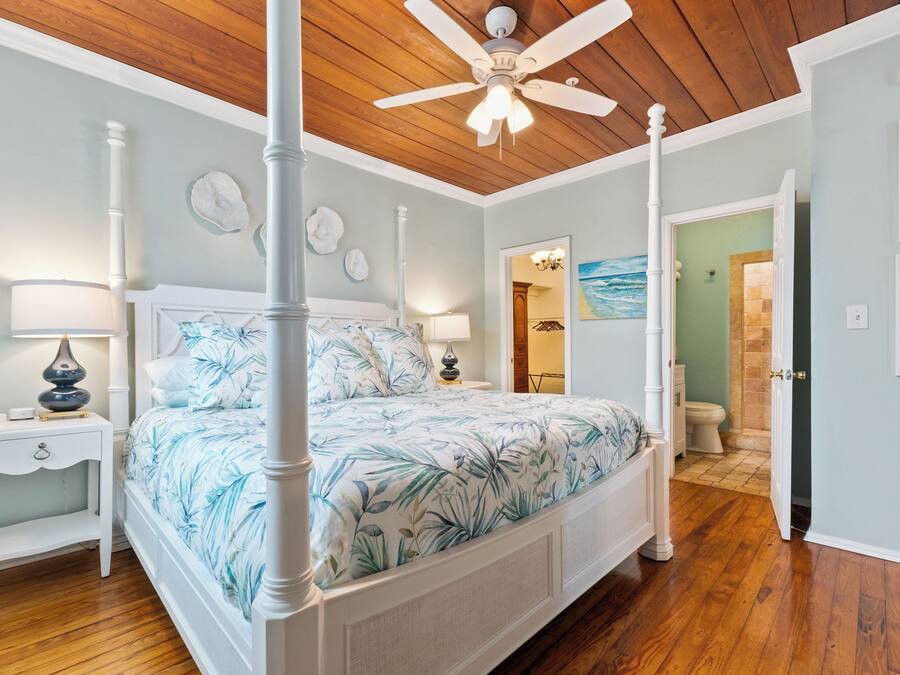 A four-poster bed with a floral bedspread, a ceiling fan, and a nightstand with a lamp.