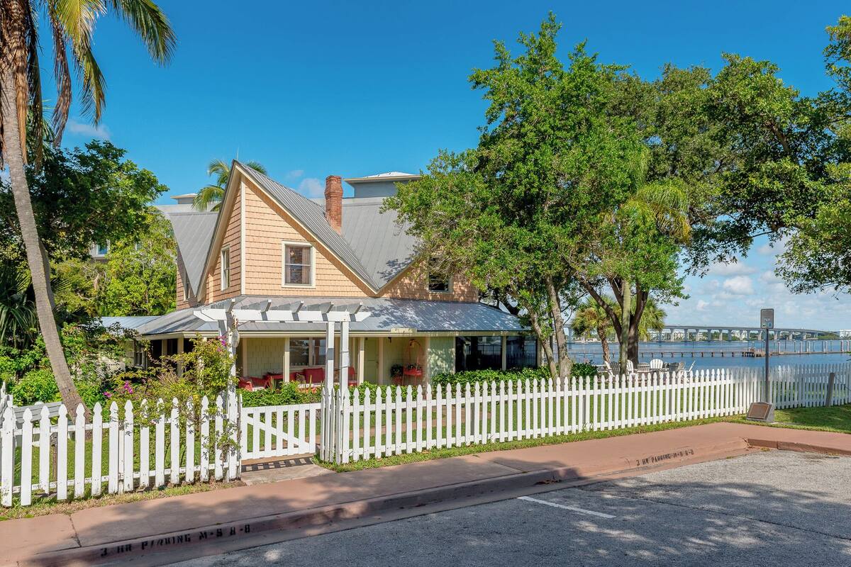 A two-story house with a porch, white picket fence, and a view of a bridge in the background.