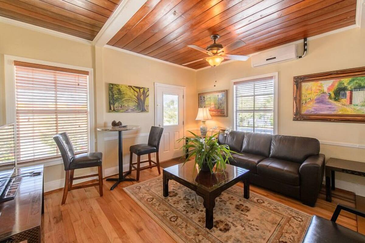 A living room with a wooden ceiling, a black leather sofa, two chairs, a coffee table with a plant, and artwork on the walls.