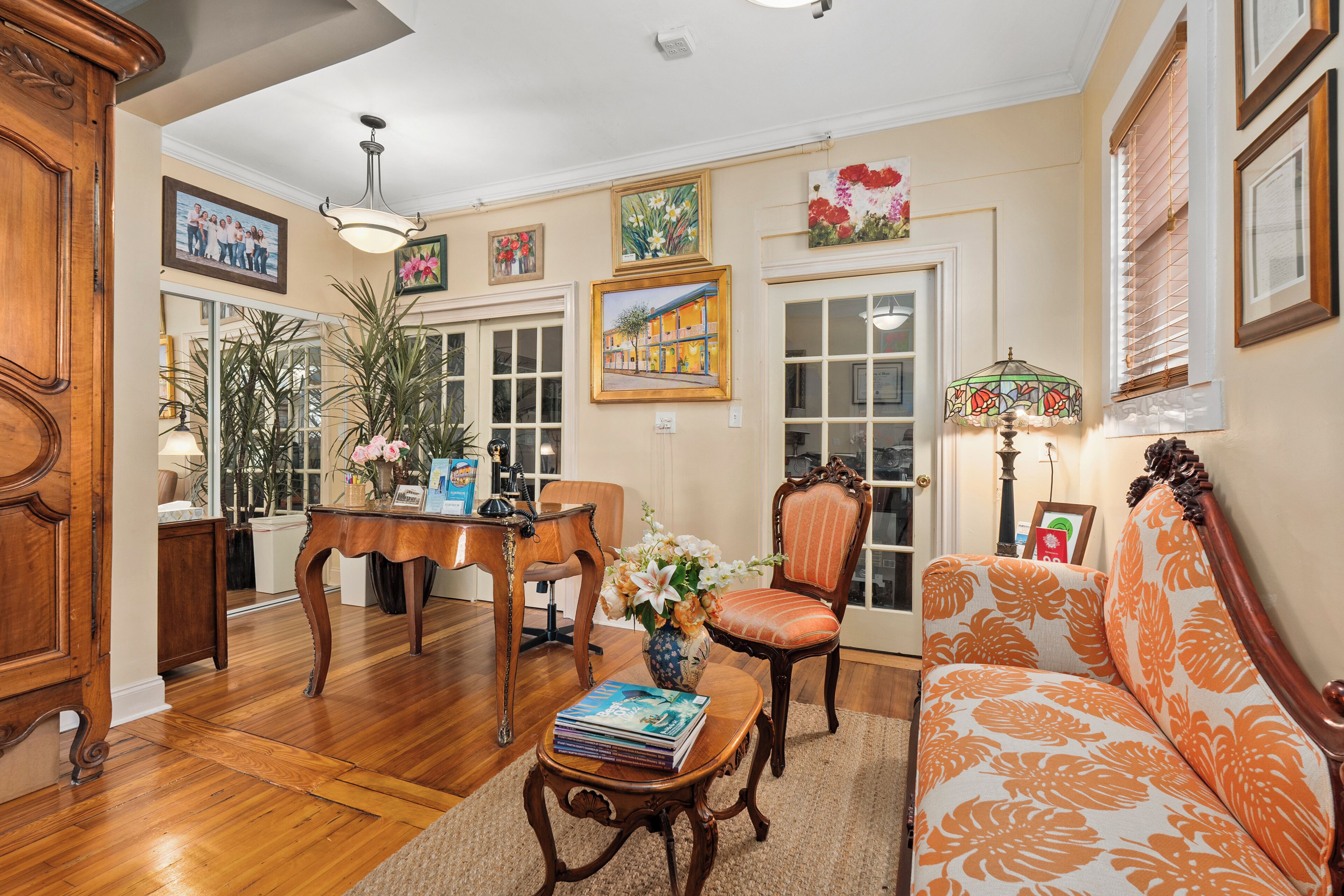 A well-lit living room with hardwood floors, a sofa, a chair, a coffee table with books, a vase with flowers, and a lamp.
