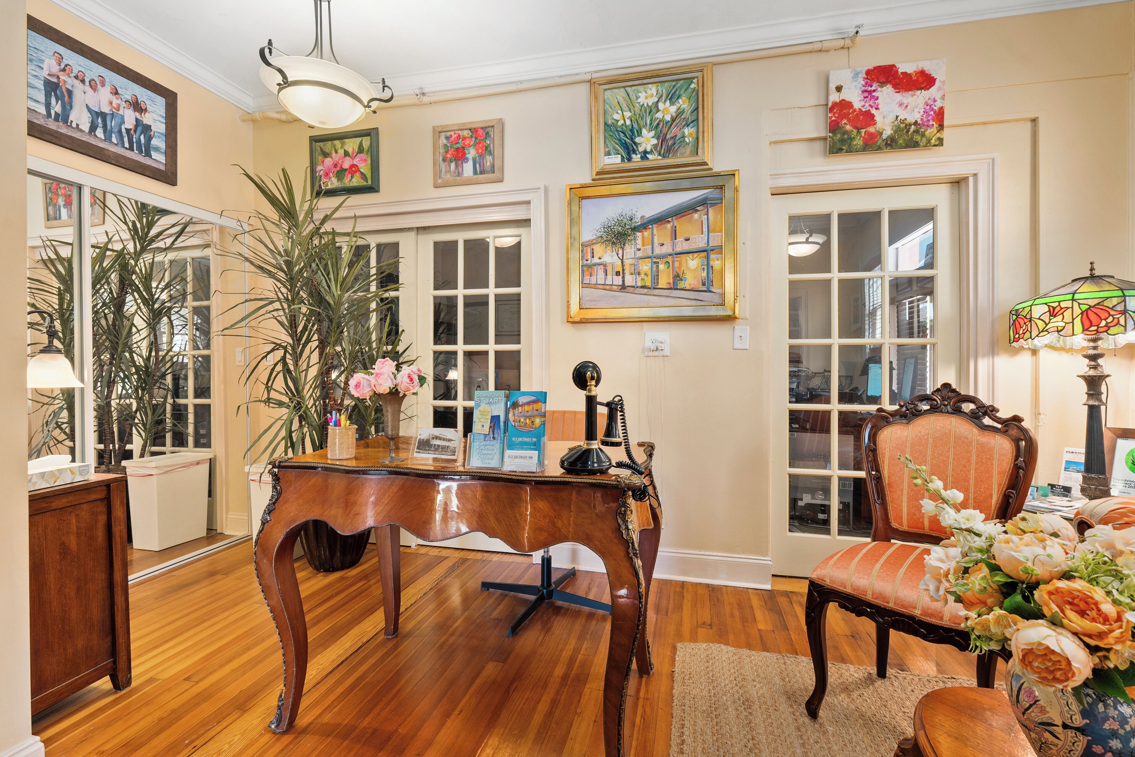 Une pièce avec un plancher en bois, un bureau en bois orné d’un vase de fleurs, une chaise avec un coussin à motifs floraux et un mur décoré d’œuvres d’art encadrées.