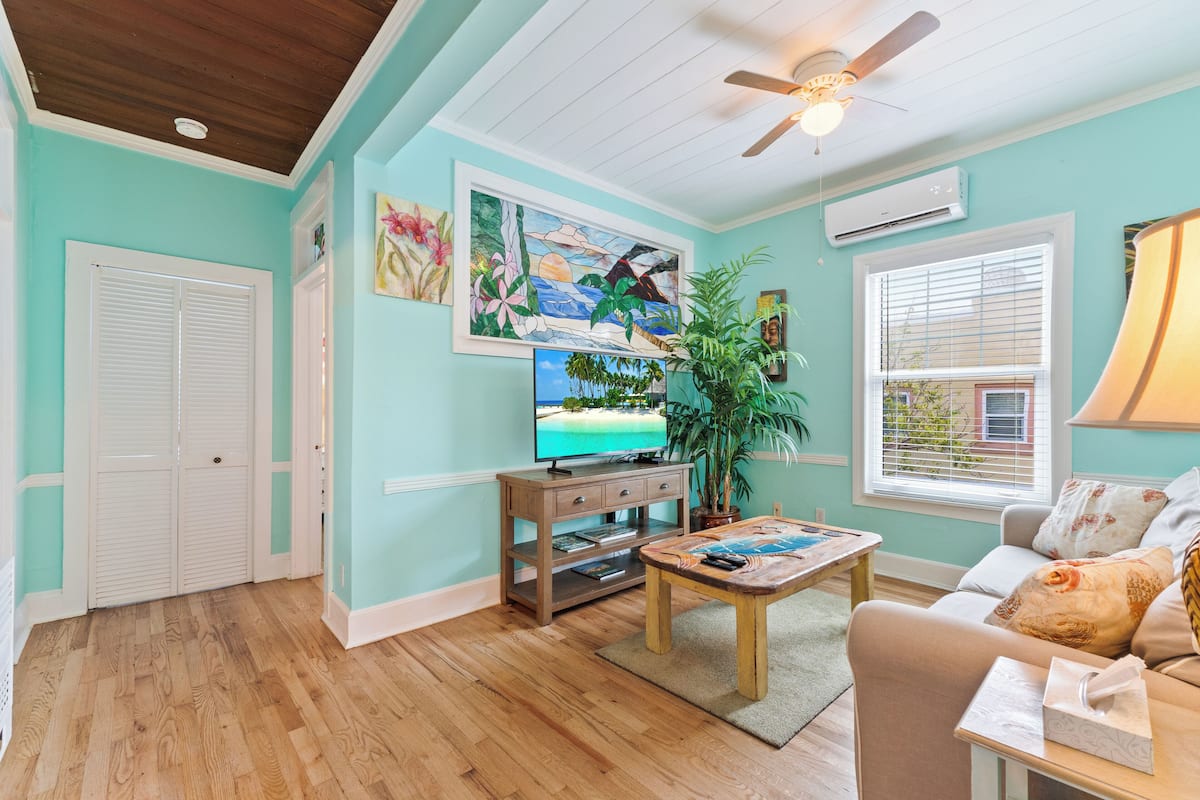 A living room with teal walls, wooden flooring, a ceiling fan, a TV on a wooden stand, a coffee table, a sofa, and a window with blinds.
