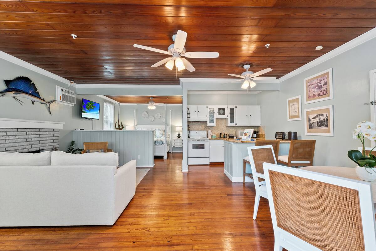 A spacious living area with a white sofa, dining table, and chairs, wooden flooring, ceiling fans, and a kitchen in the background.