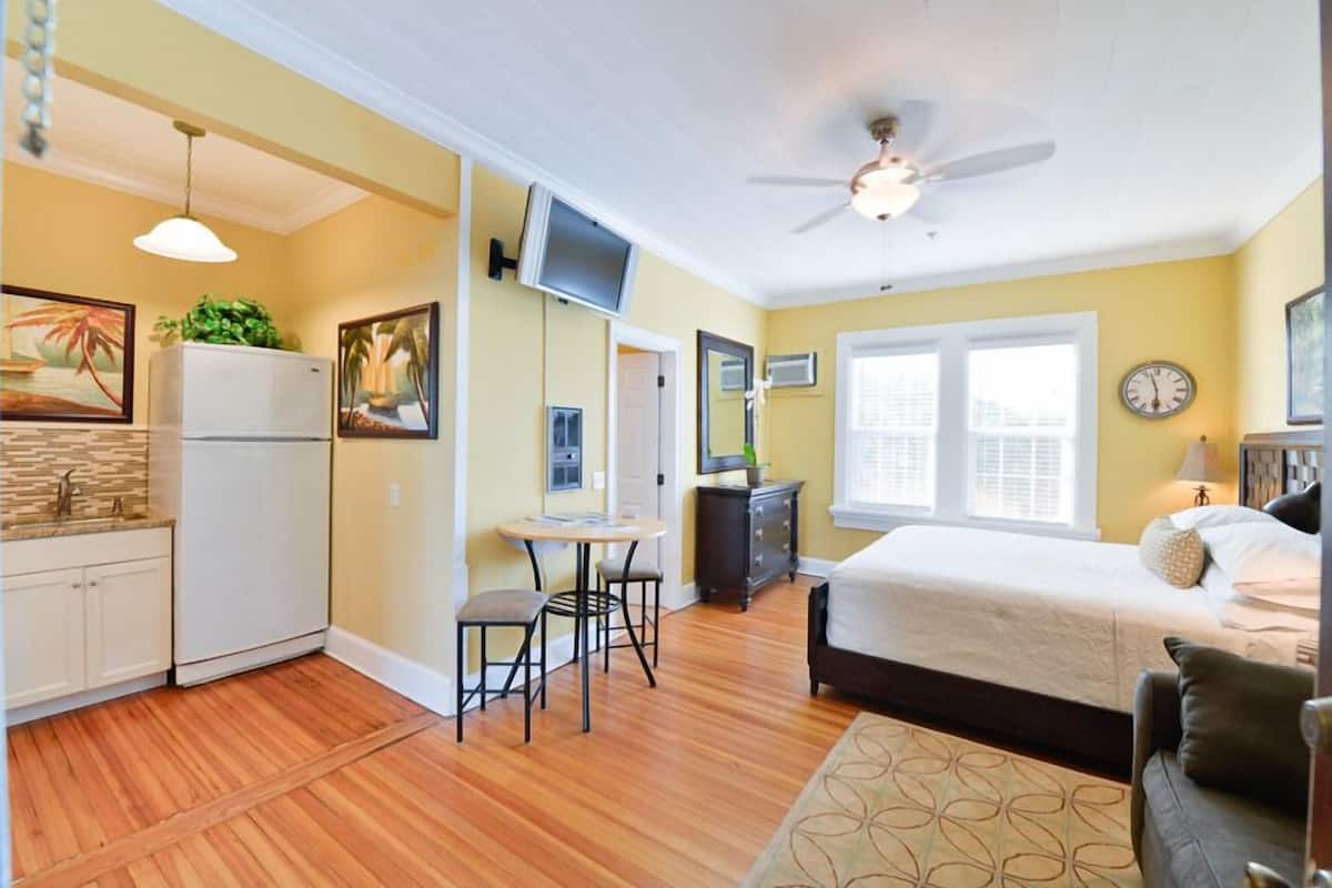 A compact hotel room with a bed, a desk, a chair, a small kitchenette, and a ceiling fan.