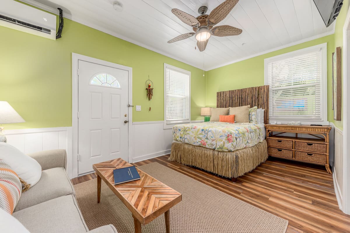 A room with a bed, a wooden table, a ceiling fan, and a window with blinds.