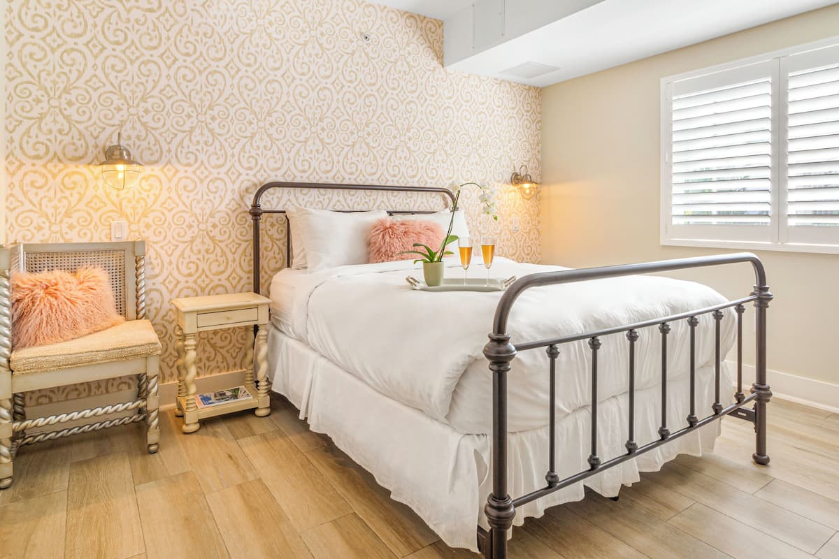 A bedroom with a bed, bedside table, chair, and a window with blinds.