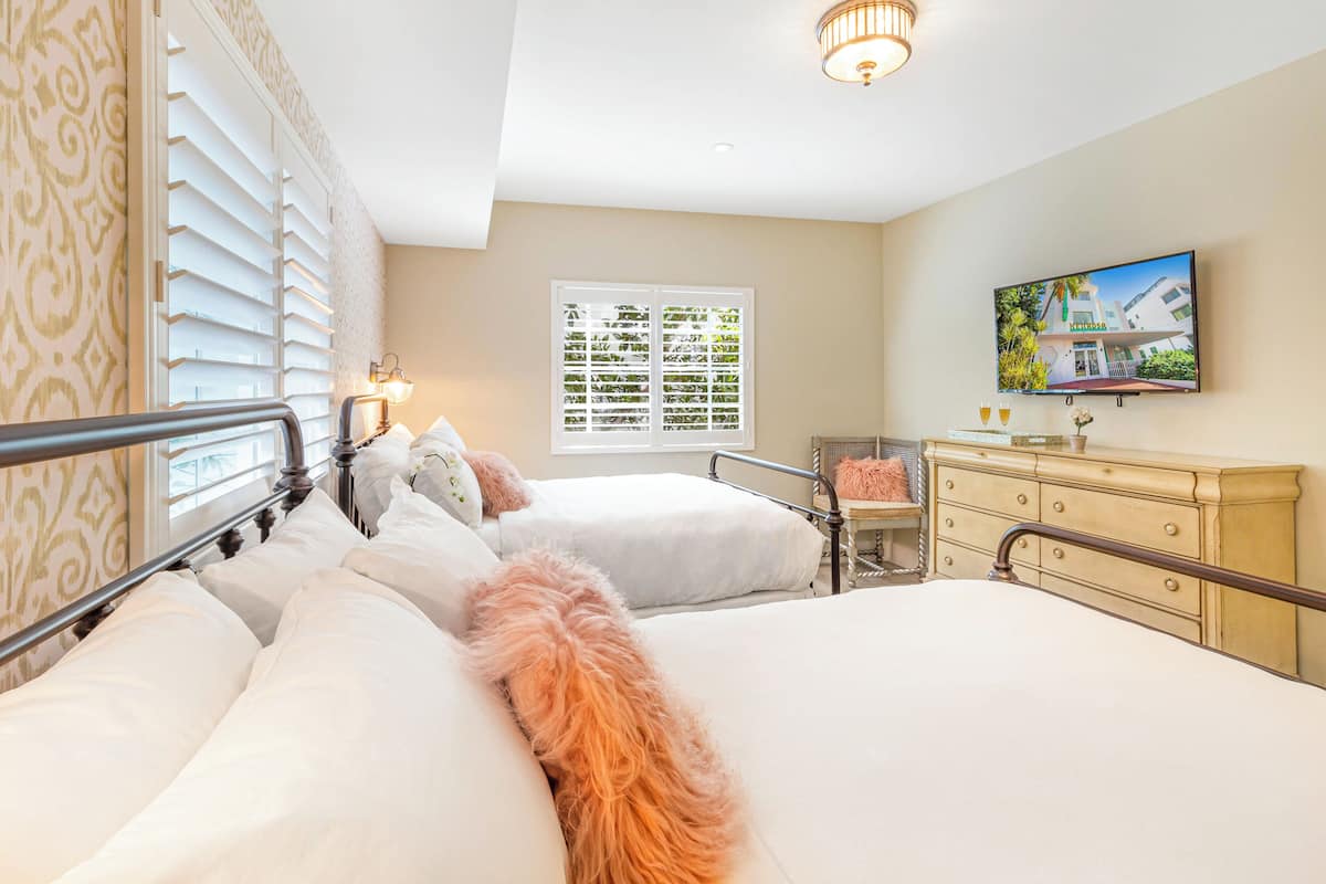 A hotel room with two beds, a television, a window with blinds, a dresser, and a lamp.