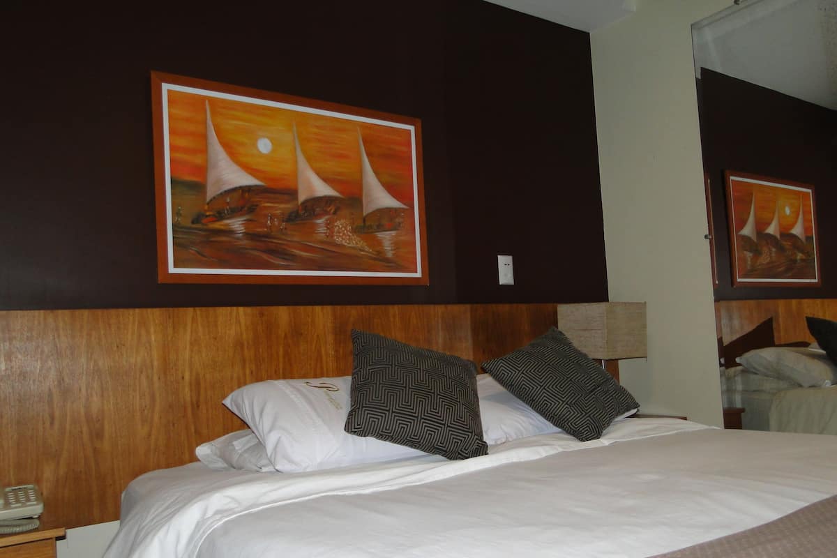 Ein Hotelzimmer mit einem großen Bett, zwei gemusterten Kissen, einem hölzernen Kopfteil, einem Nachttisch mit Telefon sowie zwei gerahmten Bildern an der Wand.
