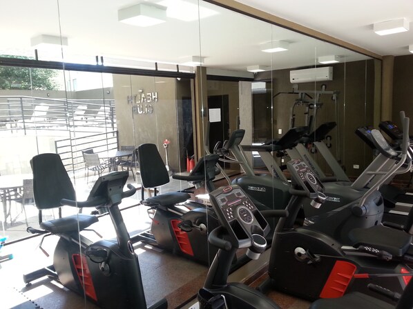 Un gimnasio con varias bicicletas estáticas y cintas de correr, amplias ventanas y un área exterior visible con mesas y sillas.