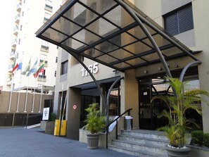 Entrada de hotel con marquesina de vidrio, escaleras y plantas en macetas.