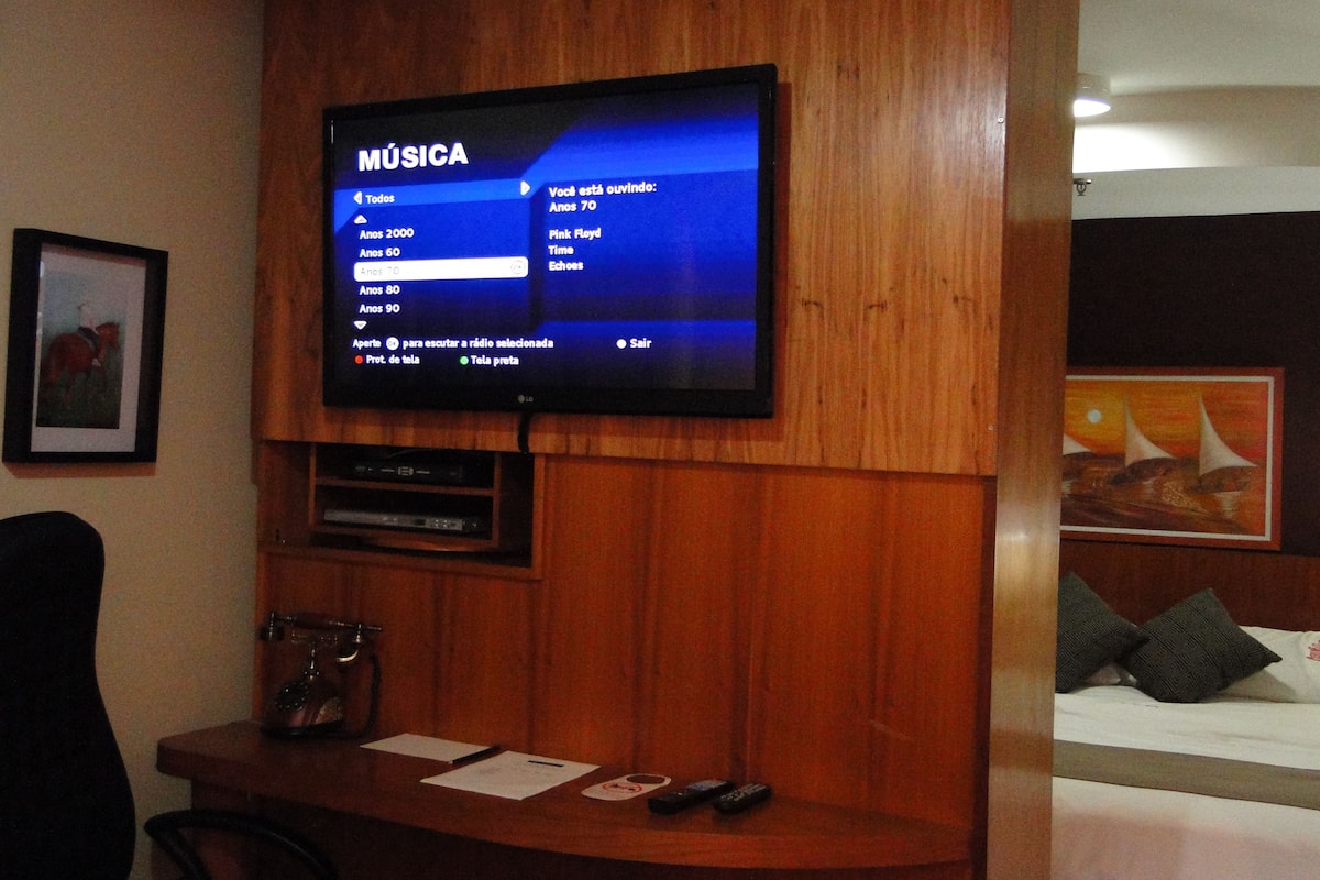 Ein Hotelzimmer mit Fernseher, der ein Musiksender-Menü anzeigt, einem Holztisch mit Kaffeemaschine und einem Bett mit Kissen sowie einem Bild an der Wand.