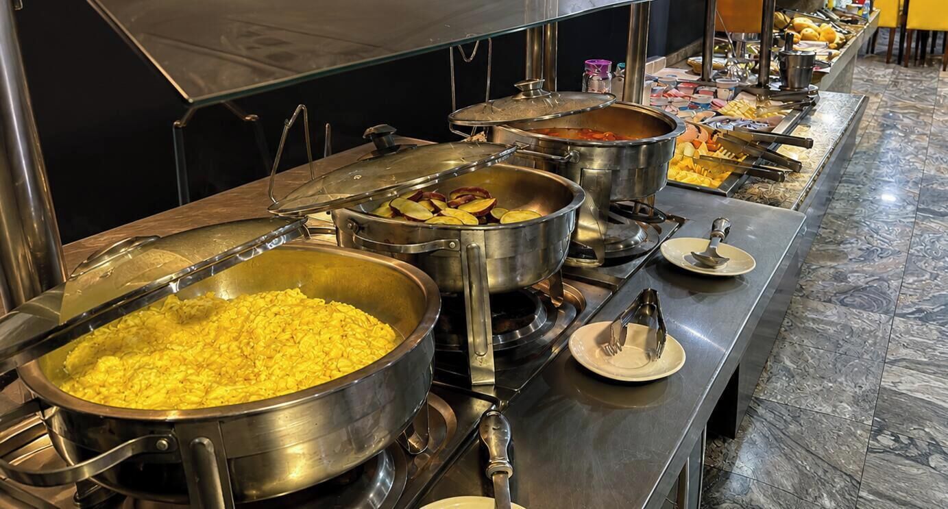 Montaje de bufé con diferentes alimentos en ollas de metal grandes, incluyendo lo que parecen huevos revueltos y un guiso.