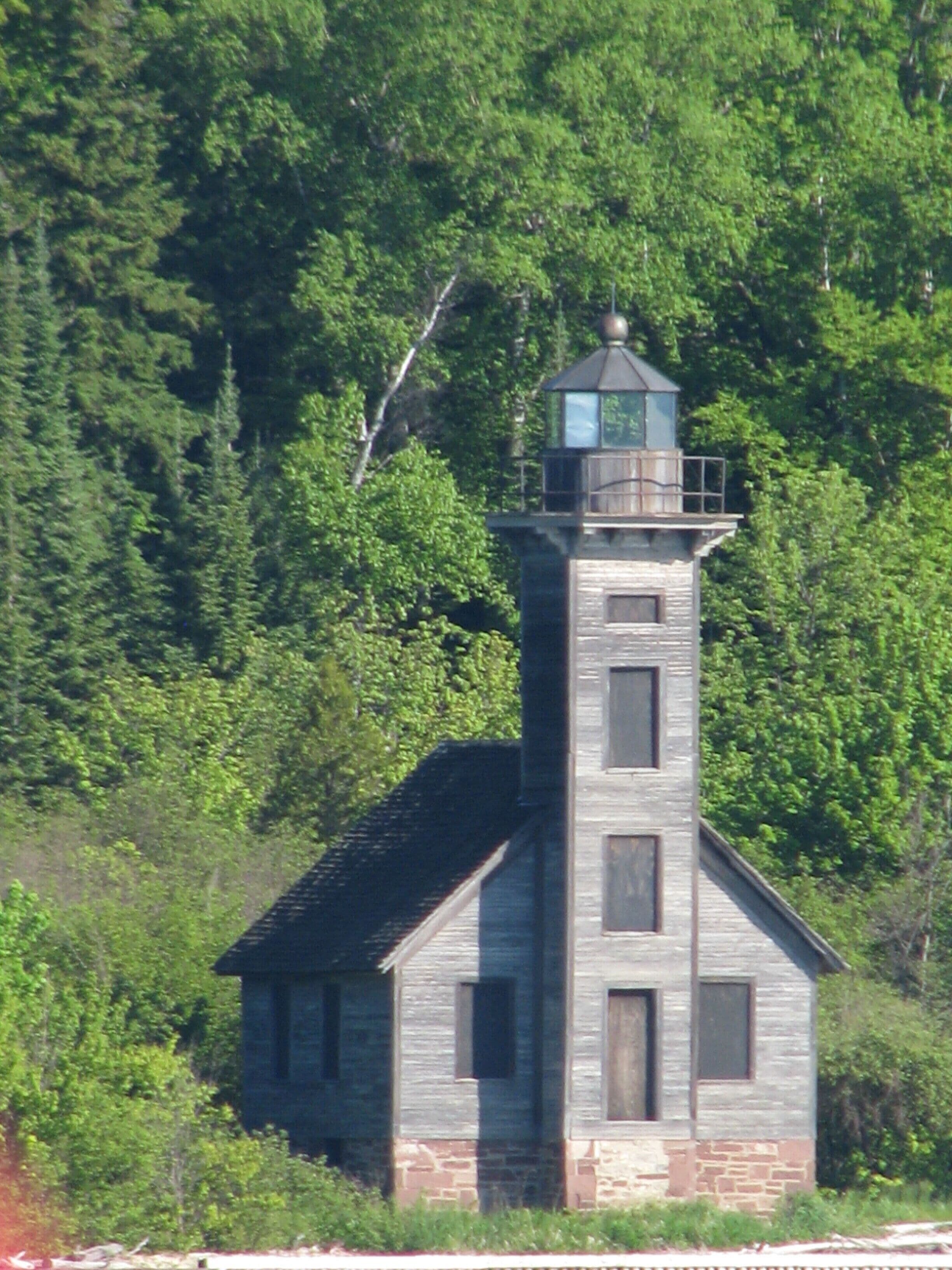 A small, rustic lighthouse with a stone base and a wooden upper structure, surrounded by dense forest.