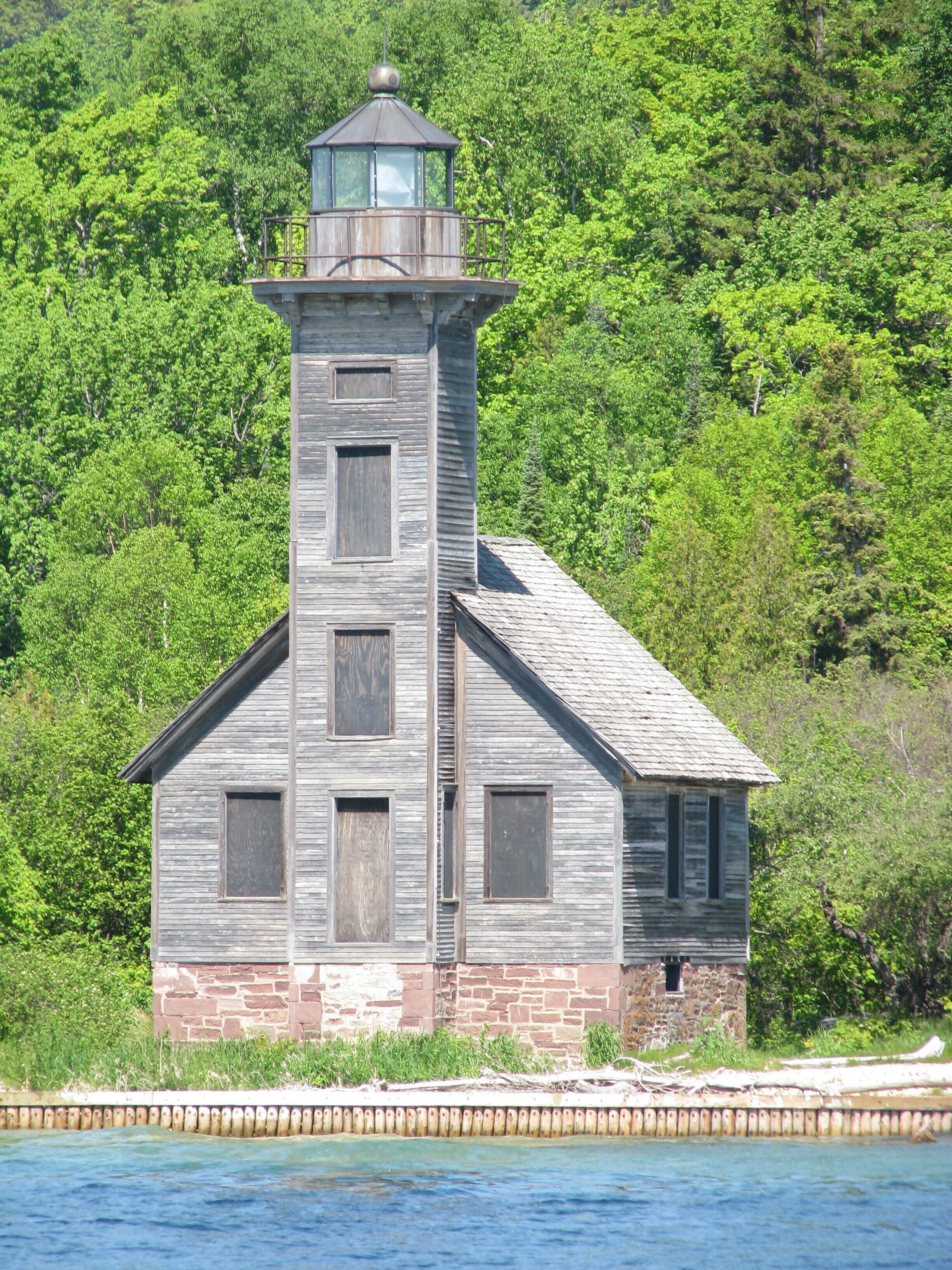 Un faro rústico con base de piedra y estructura superior de madera, rodeado de abundante vegetación.