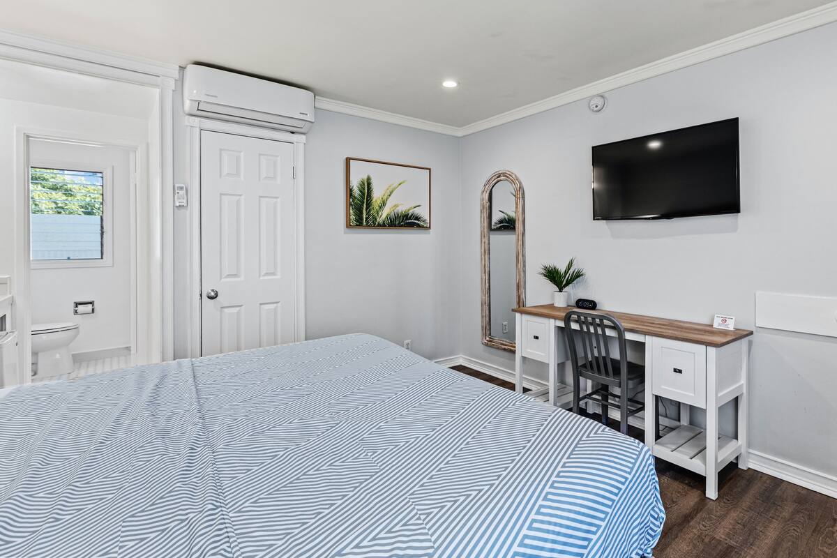 Un dormitorio con una cama, un escritorio, una silla, un espejo, un televisor y una ventana.