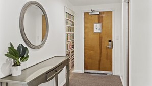 A hallway with a wooden door, a white wall, a mirror, a plant, and a bookshelf. - Ocean Lodge Santa Monica Beach Hotel (Santa Monica)