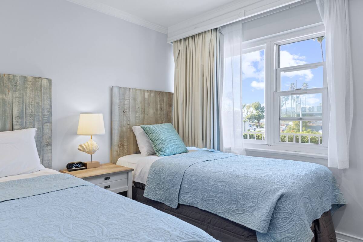 Habitación de hotel con dos camas, una mesita de noche con lámpara, un reloj y una ventana con cortinas.