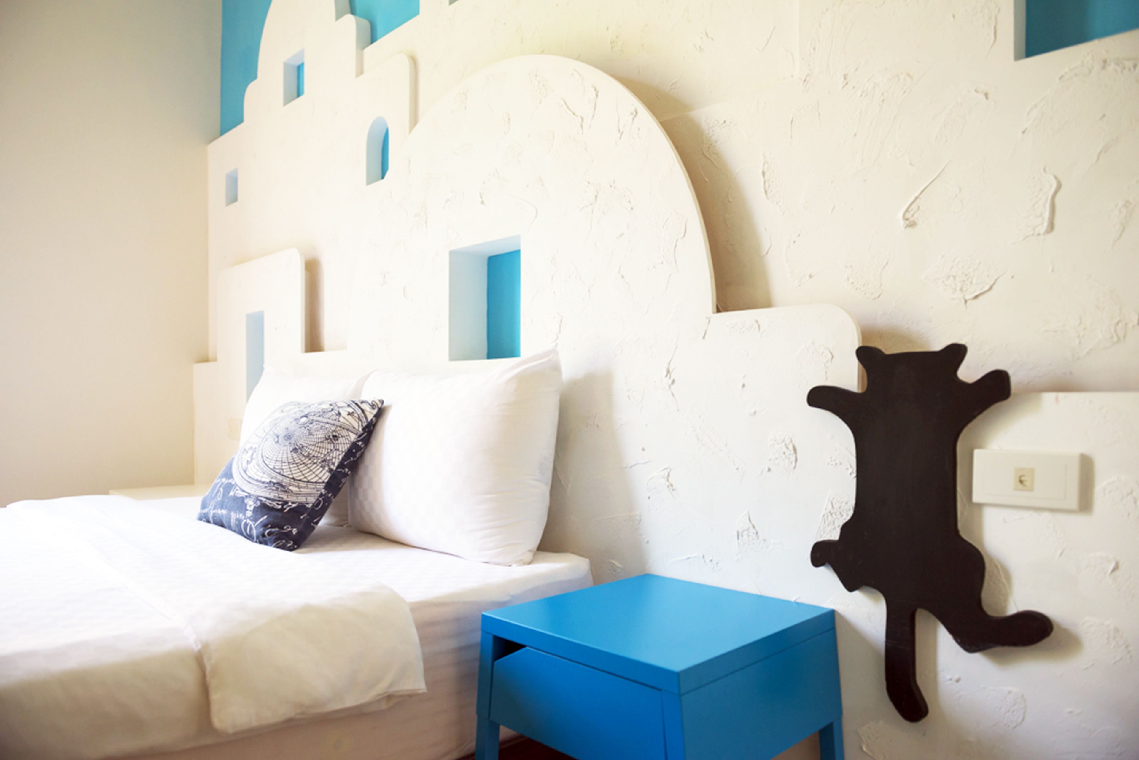 Une chambre à coucher avec un lit, une table de chevet bleue, une tête de lit blanche agrémentée de touches de bleu, et une œuvre d’art noire accrochée au mur.