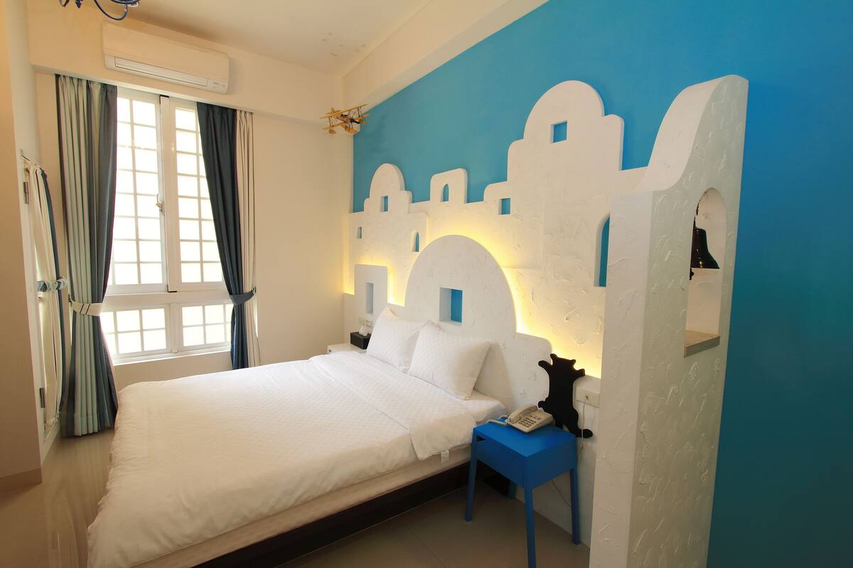 A bedroom with a bed, a nightstand with a phone, a window with curtains, and a wall mural.