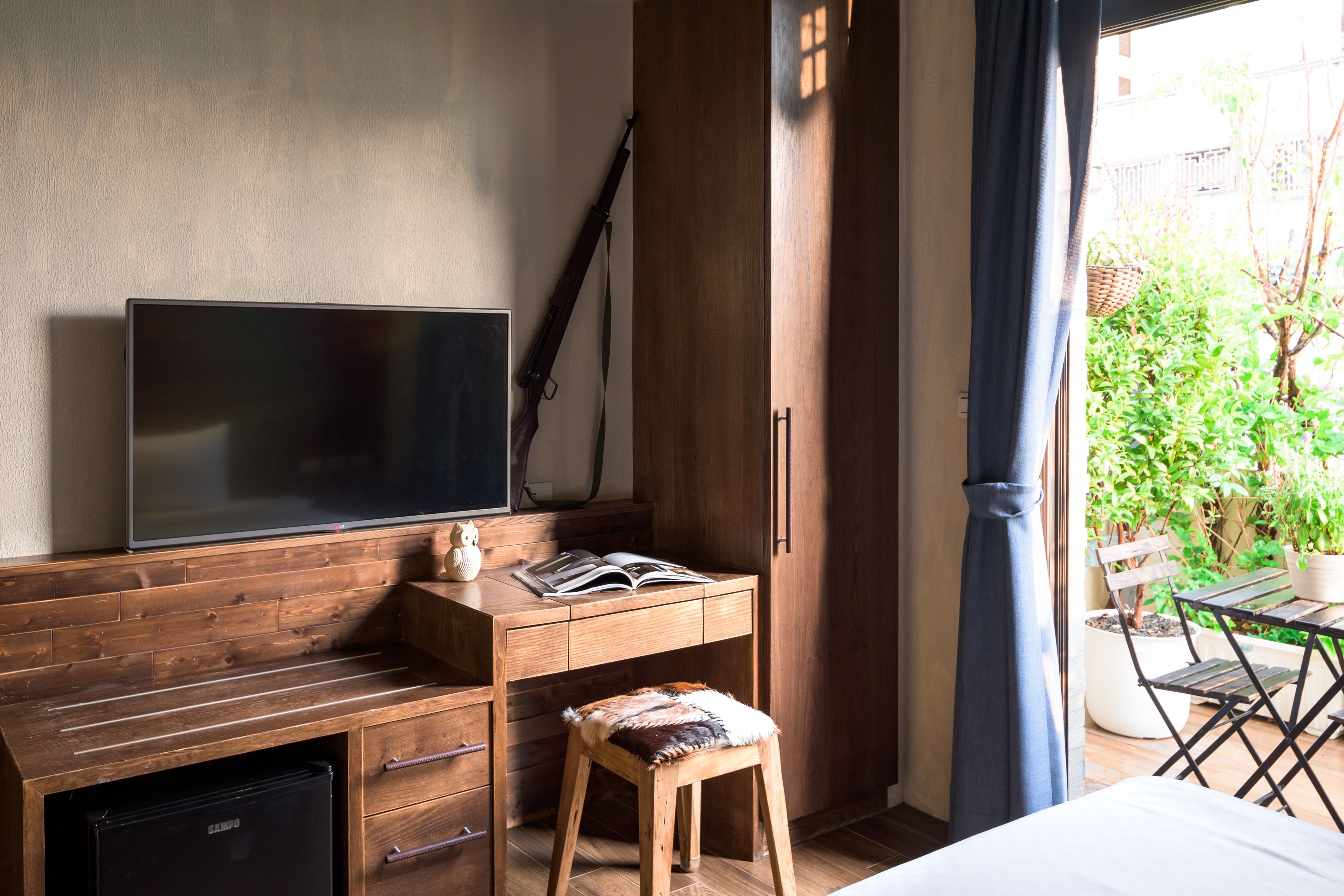 A room with a wooden desk, a television, a small stool, and a view of a garden through a window.
