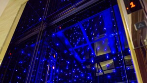 A glass elevator shaft with blue lighting and a visible emergency exit sign. - Luna's House (Kaohsiung)