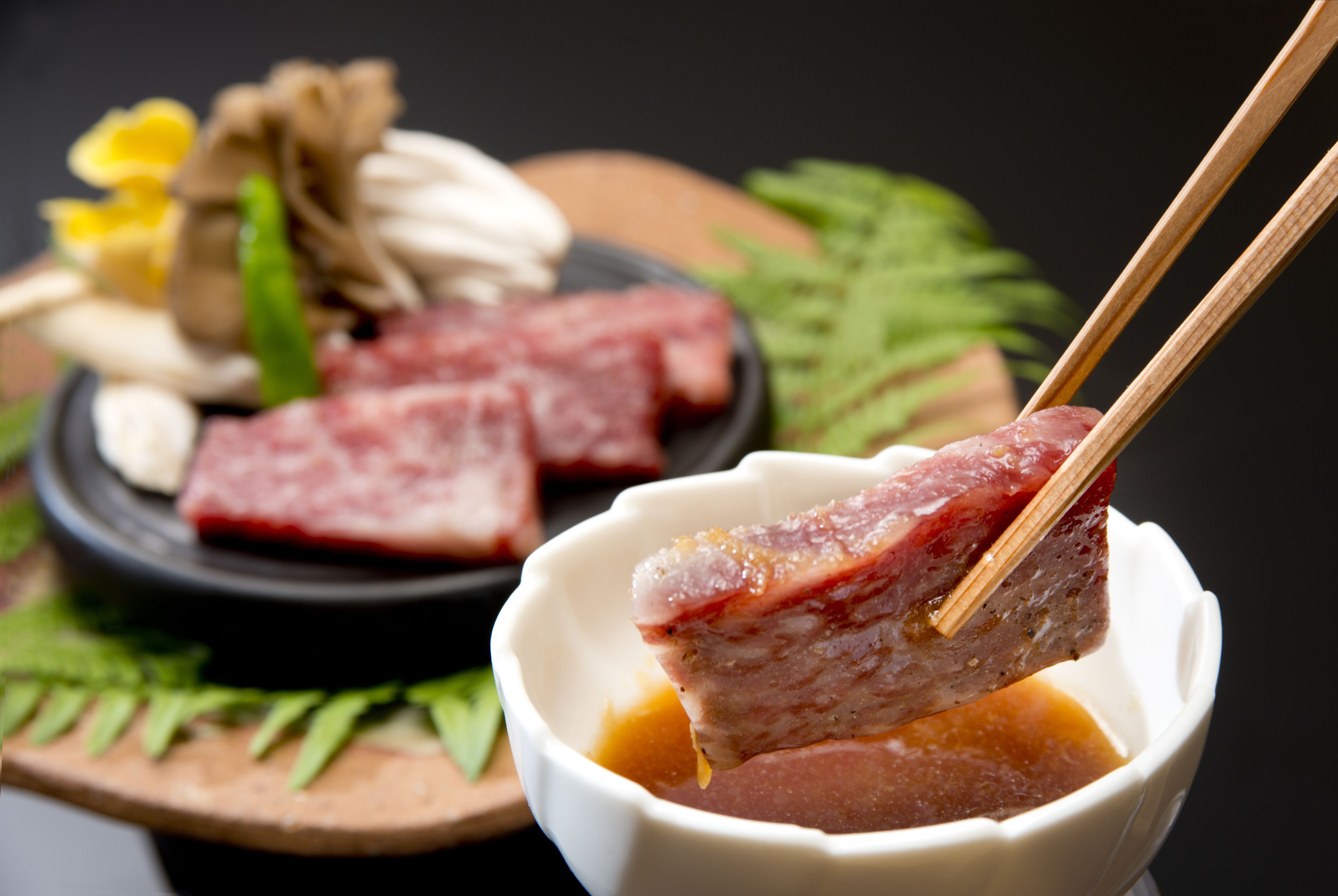 A bowl of food with chopsticks lifting a piece of meat, surrounded by other food items and garnishes on a wooden board.