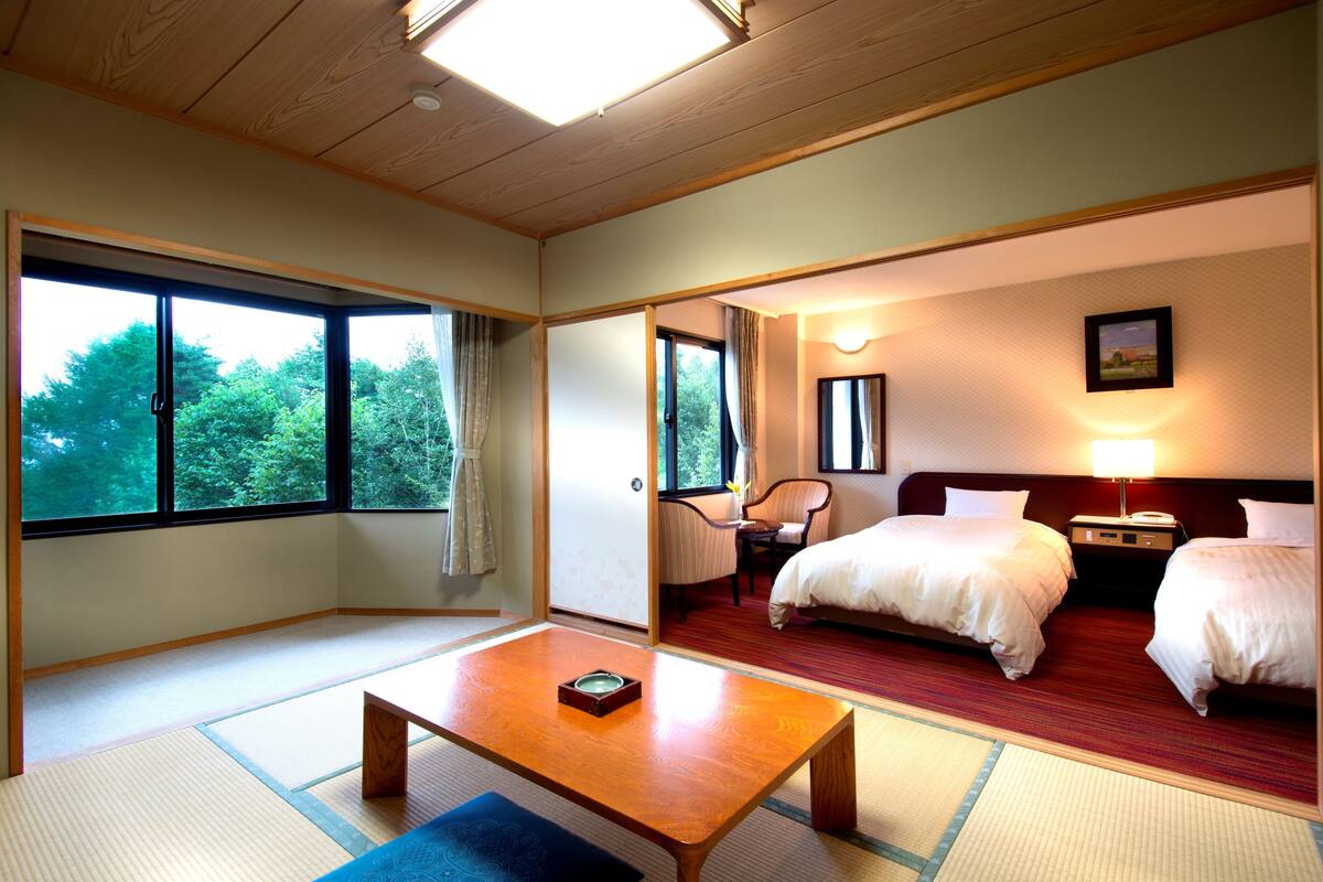 A hotel room with tatami mats, a wooden table, two beds, a chair, and a nightstand with a lamp.