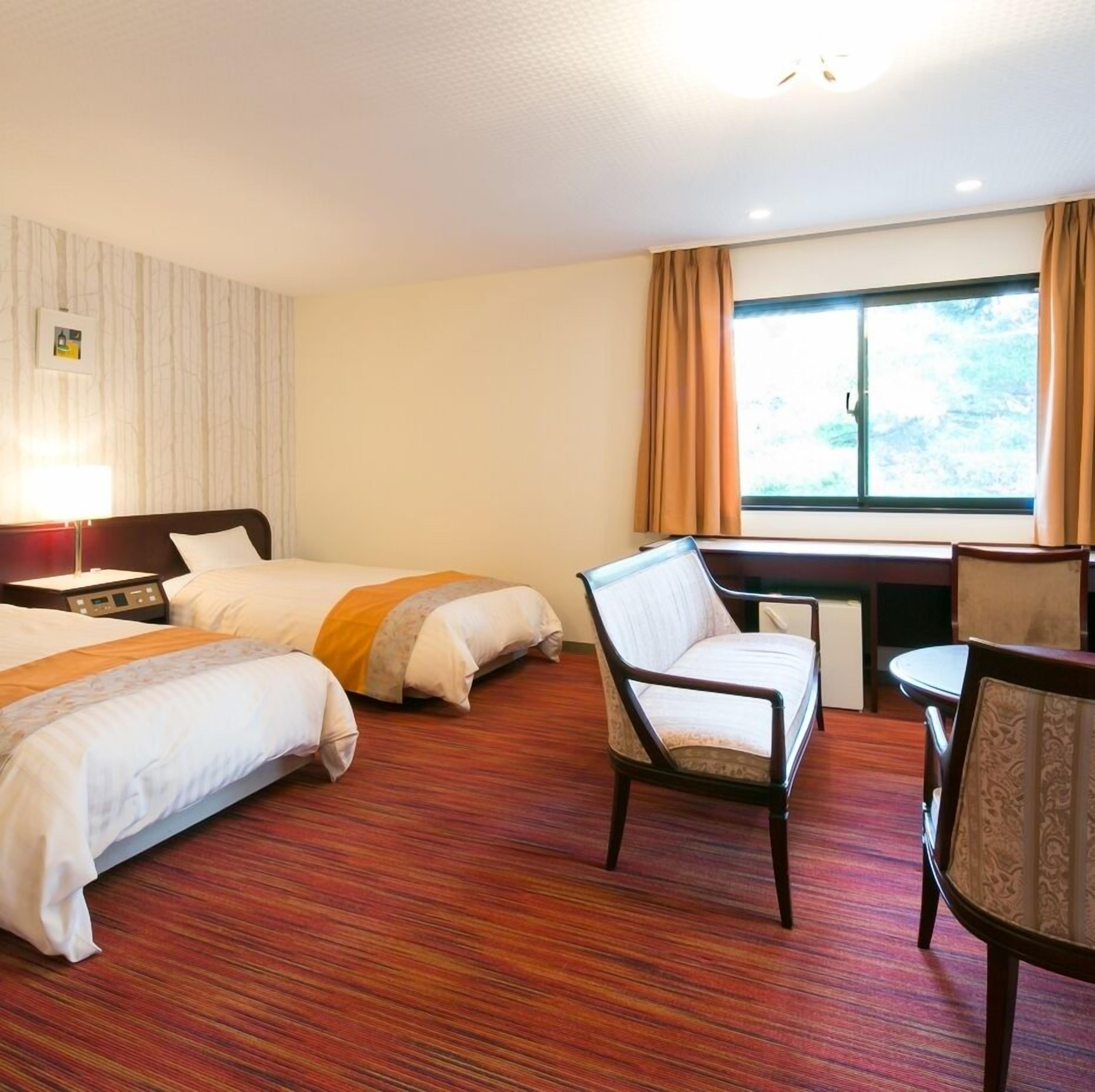 A hotel room with two beds, a desk, a chair, and a window with curtains.