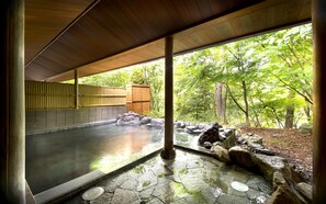 Un onsen japonés tradicional con un baño de piedra, pilares de madera y vistas a un área boscosa.