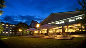 An outdoor patio area with umbrellas and chairs, a building with large windows, and a well-lit entrance. - Azumino Hotaka View Hotel (Azumino)