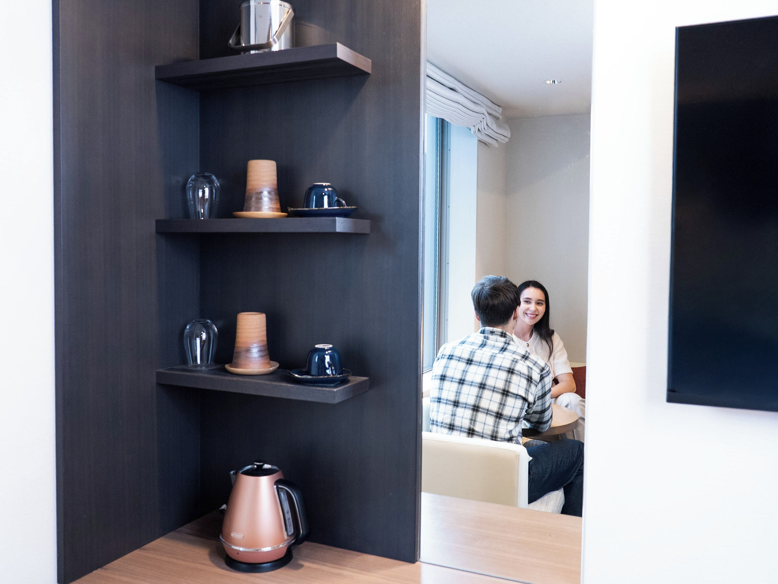 Habitación moderna con estantes de madera oscura que contienen diversos objetos, una cafetera y una vista de una pareja sentada junto a una ventana.