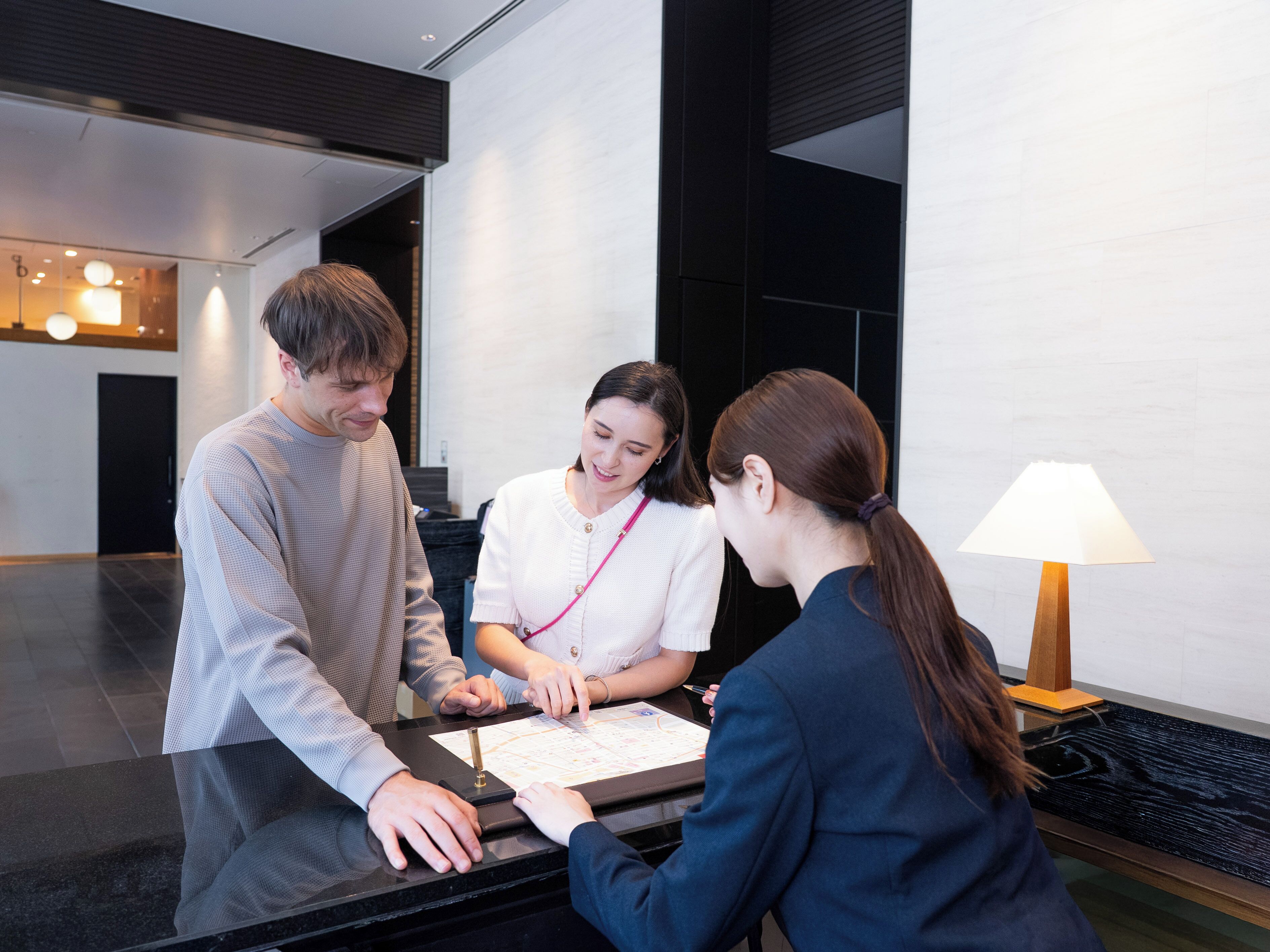 A reception desk with a person seated behind it, a standing person on the left, and another person on the right.