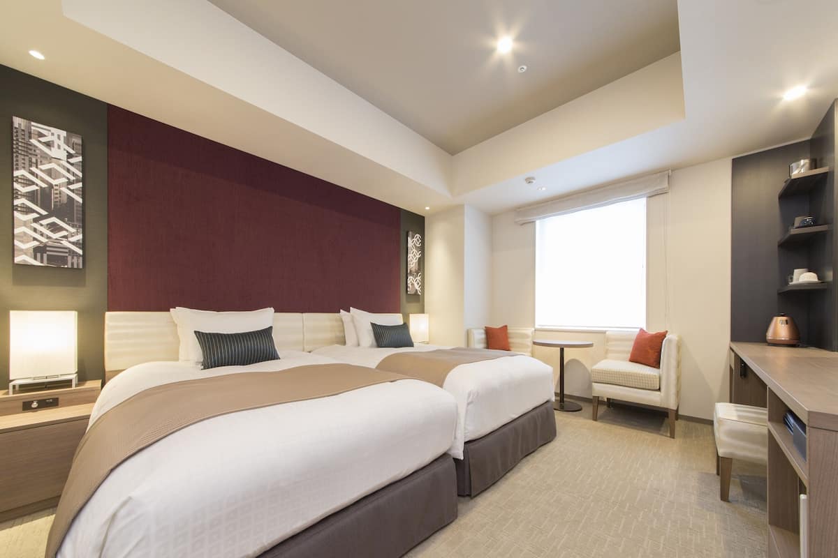 A modern hotel room with two large beds, a small seating area, and a desk.