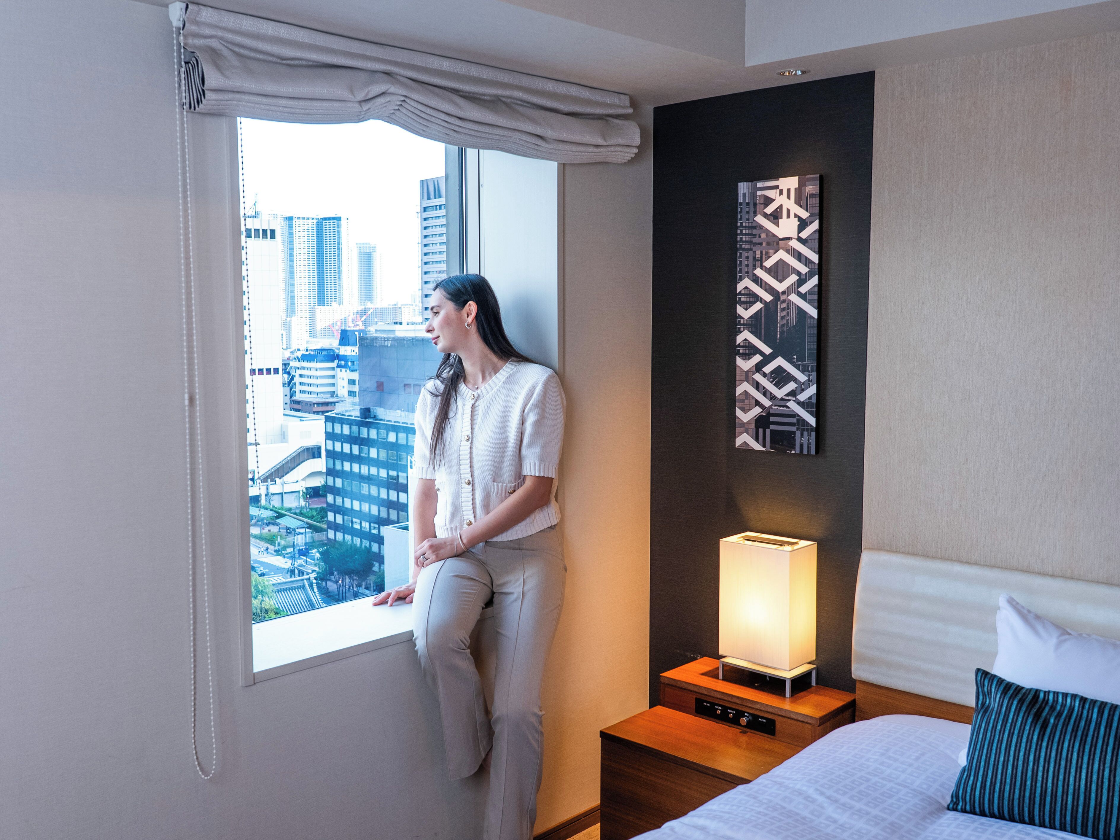 Habitación de hotel con un ventanal amplio que ofrece vistas a la ciudad, una mesita de noche con lámpara y una cama con almohada a rayas.