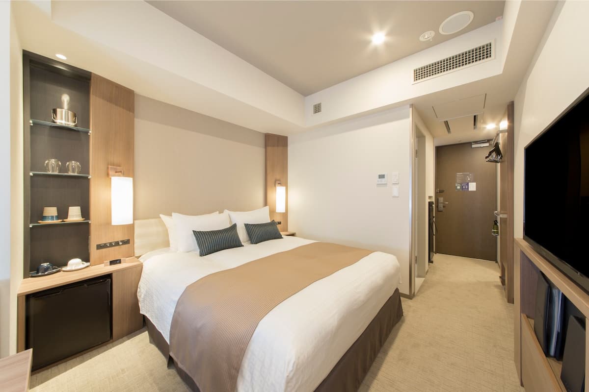 A hotel room with a large bed, bedside tables, a built-in TV, a mini-fridge, and a shelf with glasses and a hat.