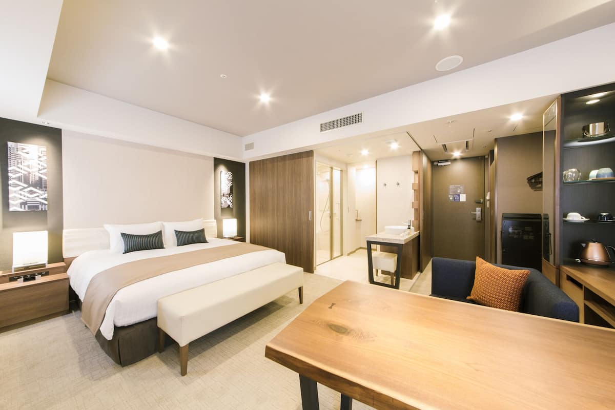 A modern hotel room with a large bed, bedside table, and a wooden bench. There is a small table with a chair and a shelf with kitchen items.