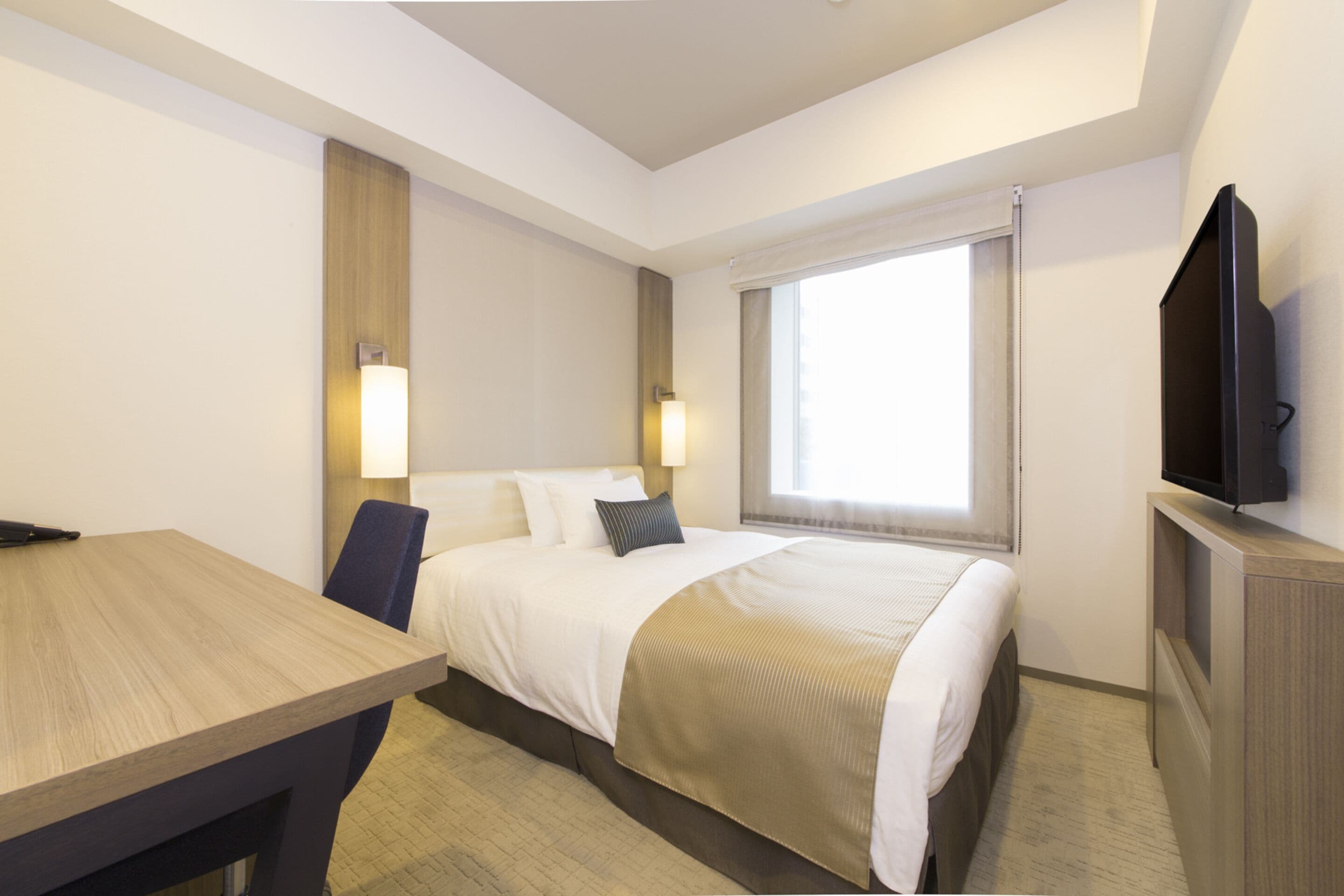 A hotel room with a bed, bedside lamps, a TV, a desk, and a window with curtains.
