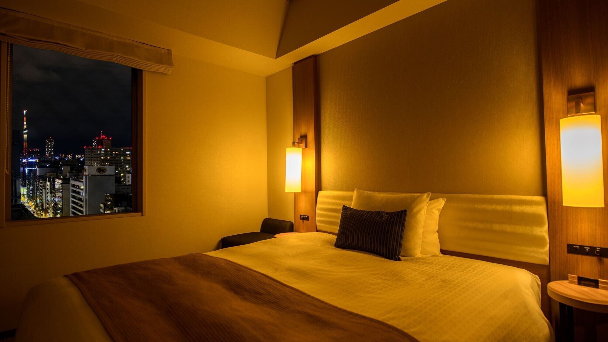 A hotel room with a large bed, two bedside tables, a nightstand, a window with a city view, and a wall-mounted light fixture.