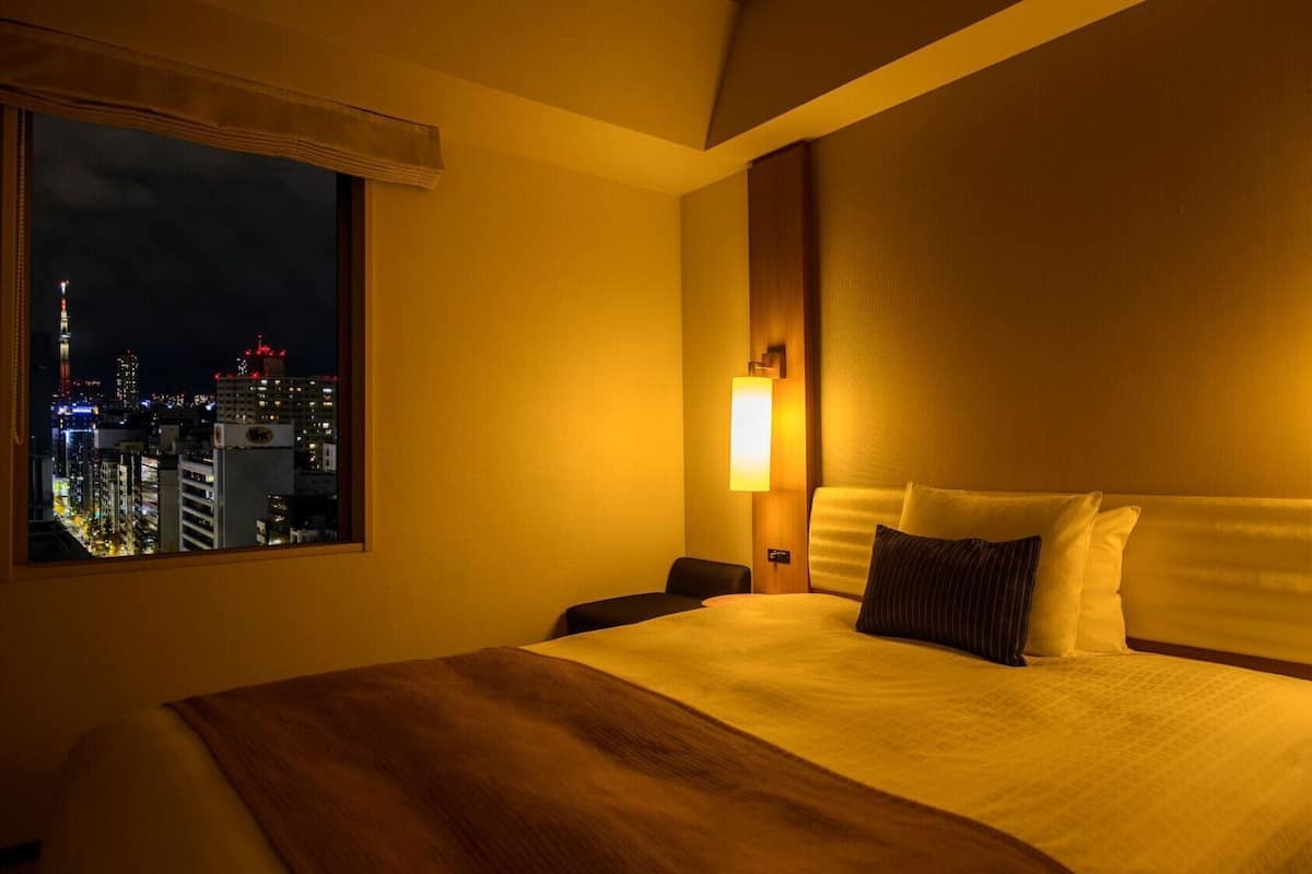 A hotel room with a large bed, two bedside tables, a nightstand, a window with a city view, and a wall-mounted light fixture.
