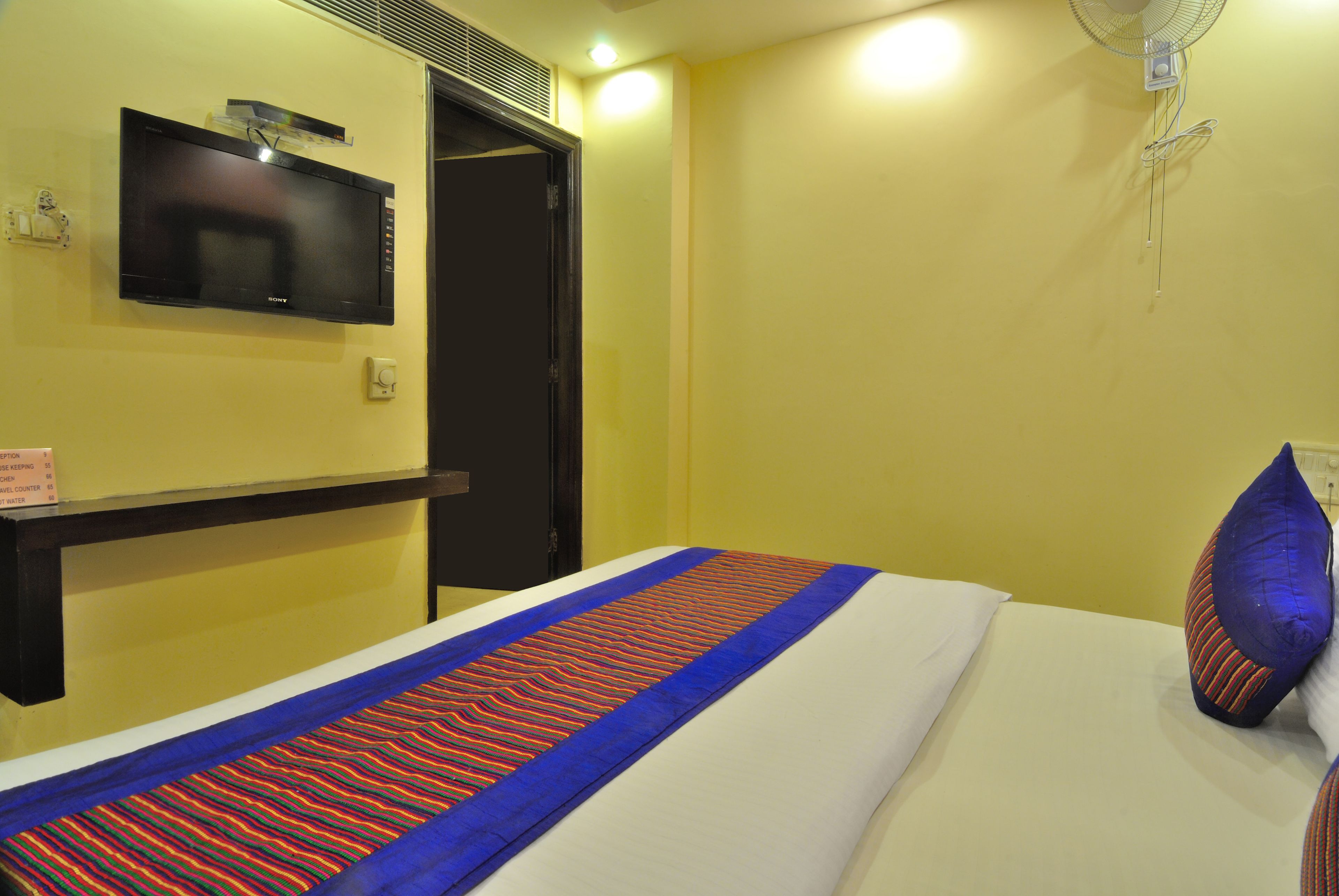 Una habitación de hotel con una cama, un televisor de pantalla plana montado en la pared, una mesita de noche y un ventilador de pared.