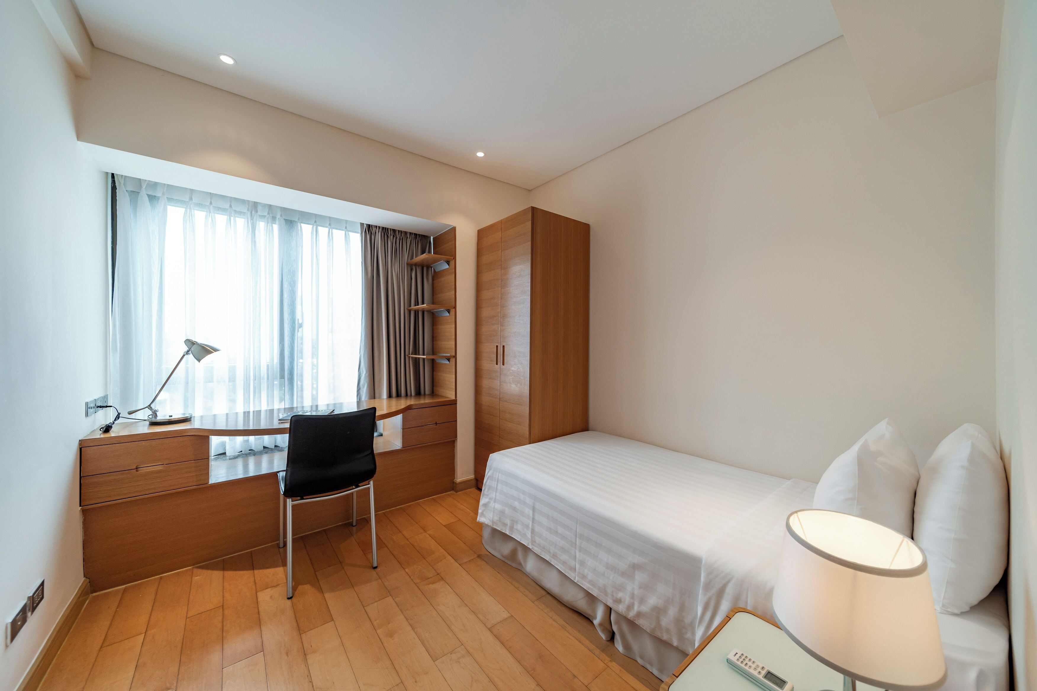 A compact hotel room with a wooden desk, a single bed, a chair, and a bedside table with a lamp.