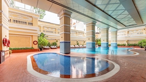 A large indoor swimming pool with tiled flooring and a glass ceiling, surrounded by columns and potted plants.