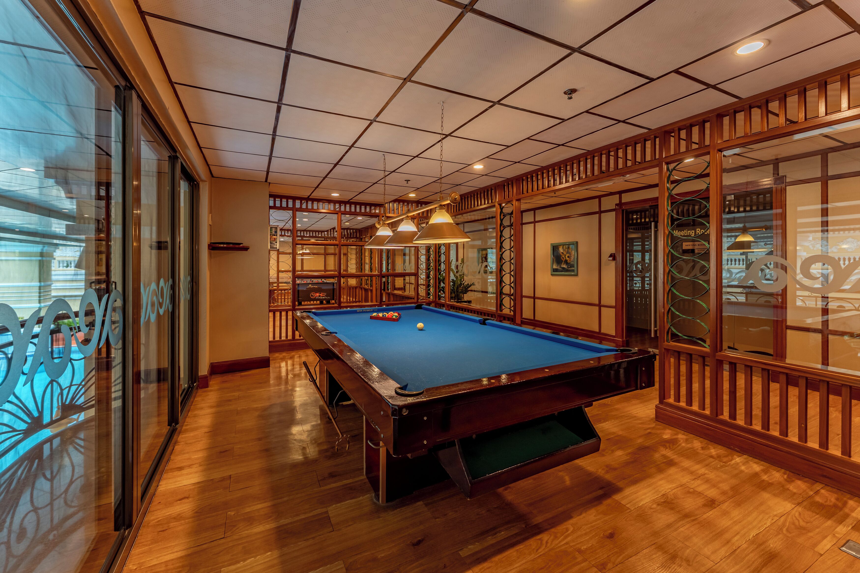 Una mesa de billar en una sala con suelo de madera y paredes de vidrio, con un techo que presenta un diseño de cuadrícula y lámparas colgantes.