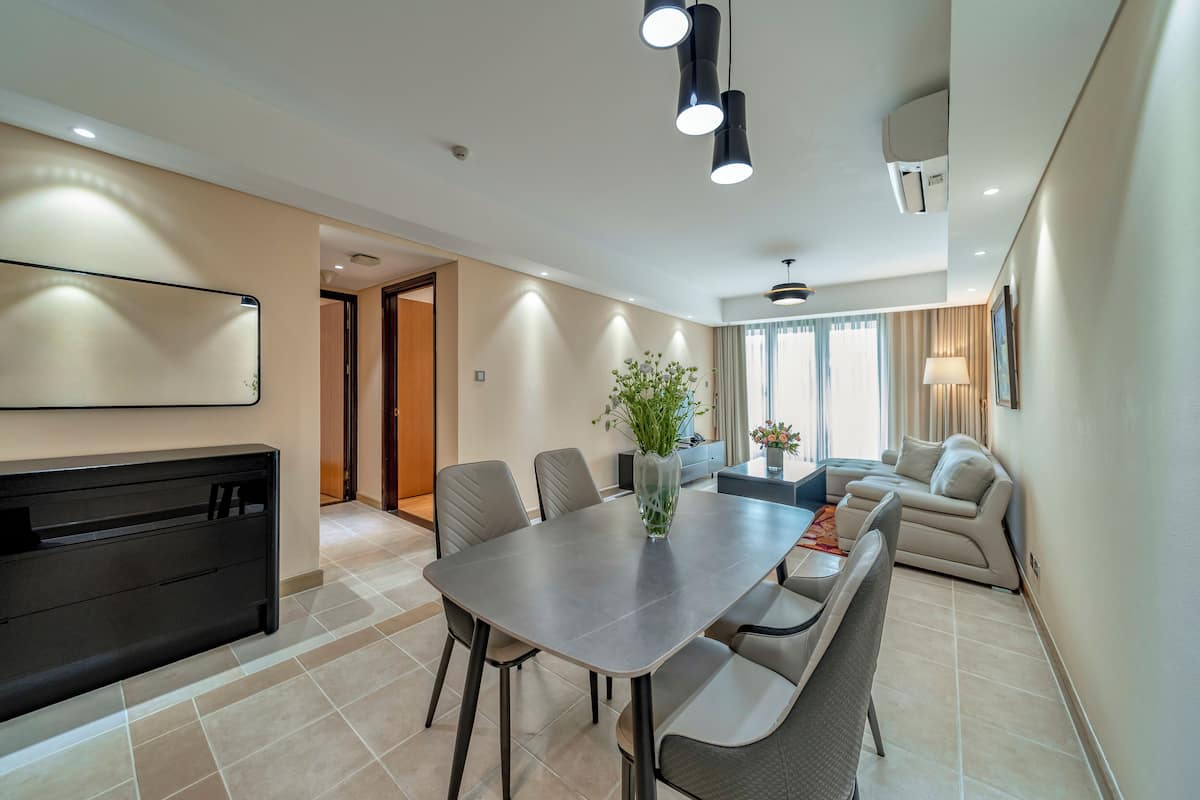 A modern dining area with a rectangular table, multiple chairs, a vase with greenery, and a sofa in the background.