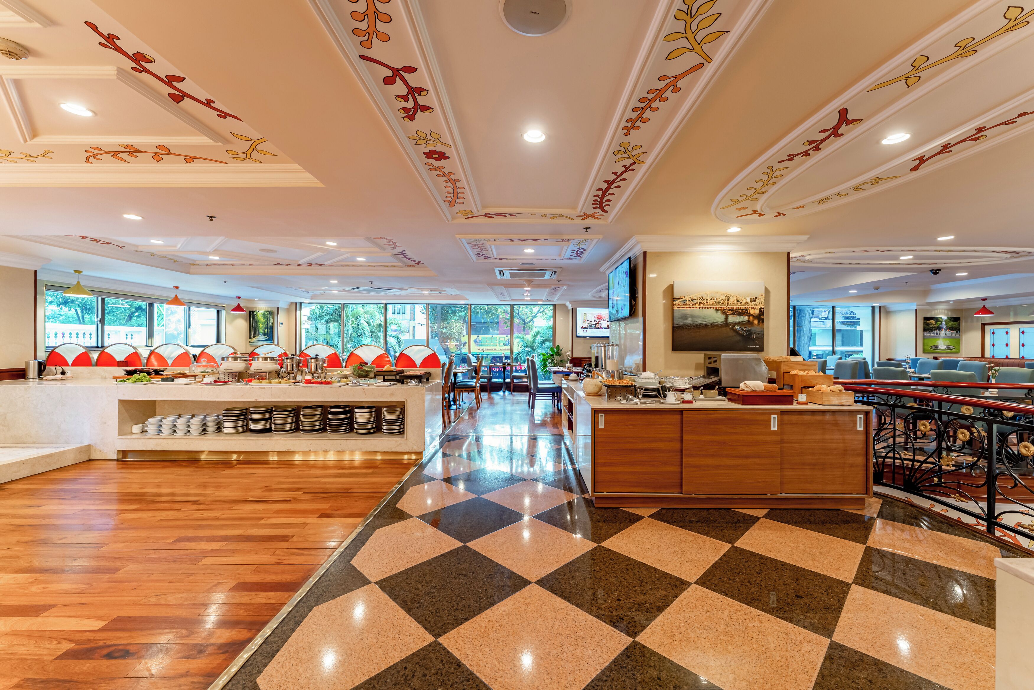 A spacious hotel lobby with a checkered floor, a central reception desk, and a dining area with red chairs.