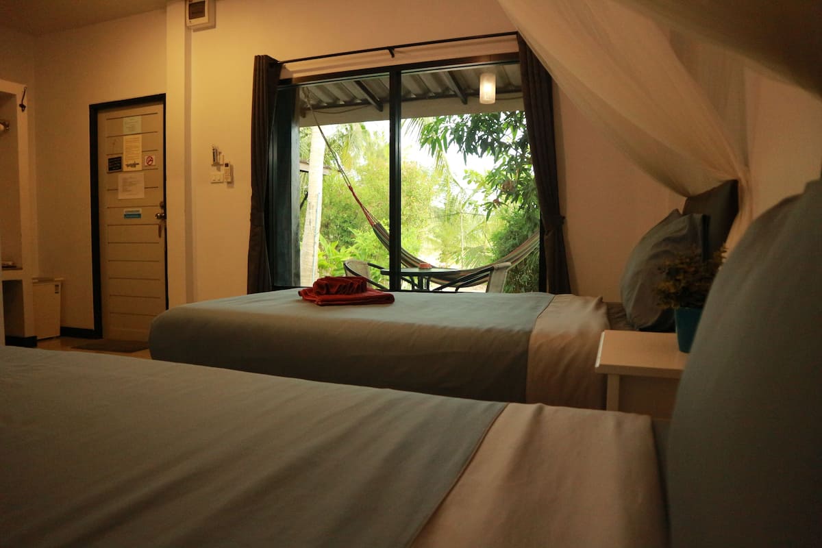 Un dormitorio con dos camas, una ventana con vistas a zonas verdes, una puerta con un letrero y una mesita con una planta.