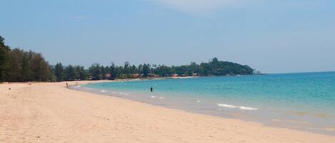 A wide sandy beach with clear blue water, a few people, and a forested hill in the background.