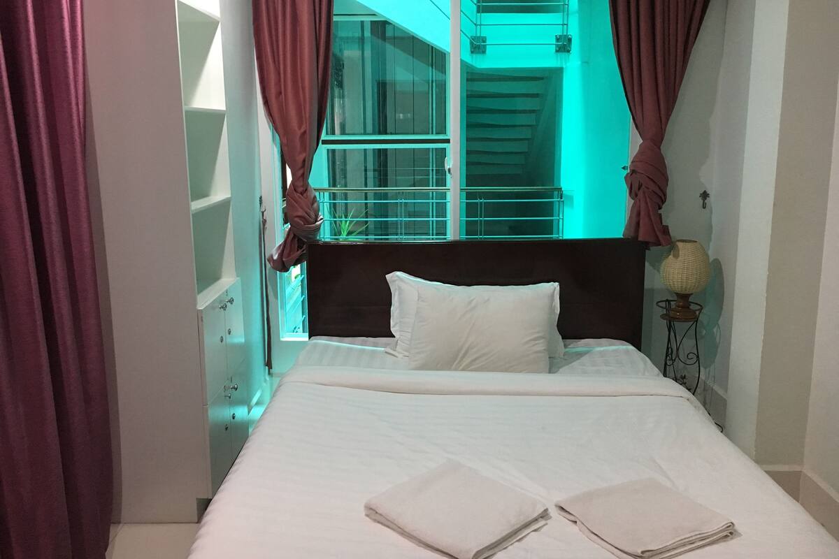 Una habitación de hotel con cama, mesita de noche, lámpara y ventana con cortinas.