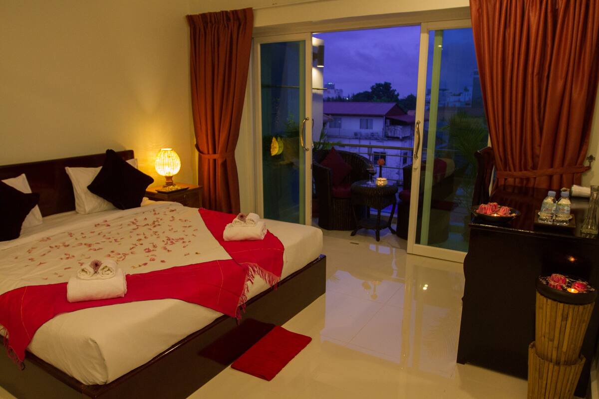Habitación de hotel con una cama grande, una mesita de noche con lámpara, un balcón con mesa y sillas, y vistas al exterior.