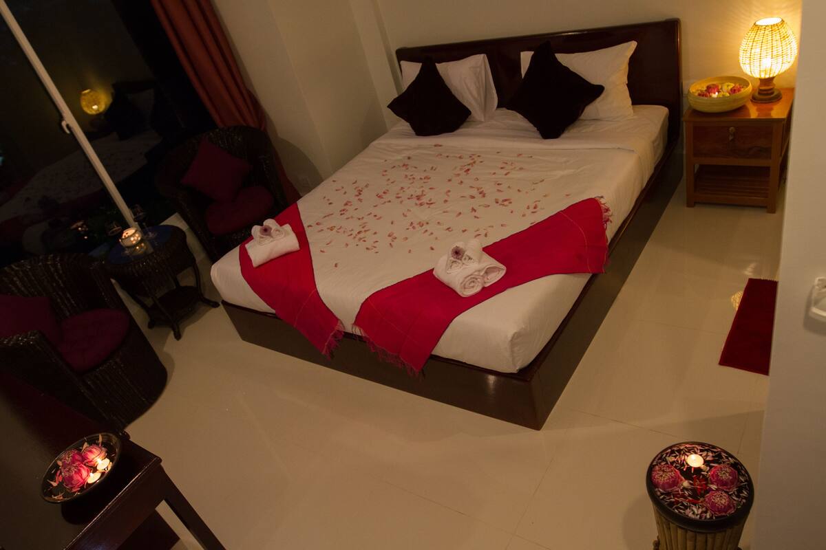 Una cama bien hecha con una manta roja y almohadas blancas, una mesita de noche con una lámpara, y una pequeña mesa con una vela.